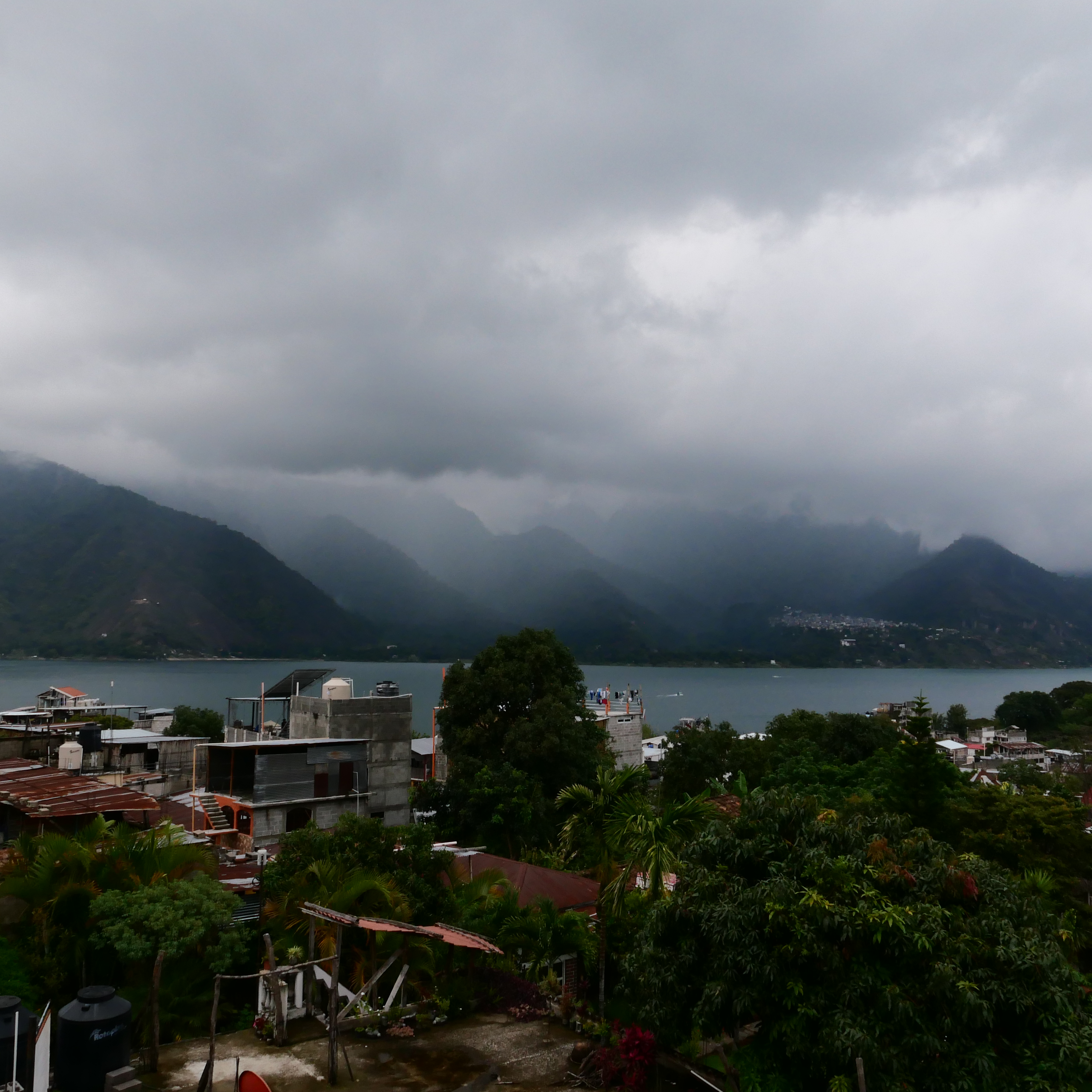 A photo from a balcony in a Lake Atitlan town, of the skyline with the buildings in front of them, the lake, and the mountains, with layered clouds covering much of them.