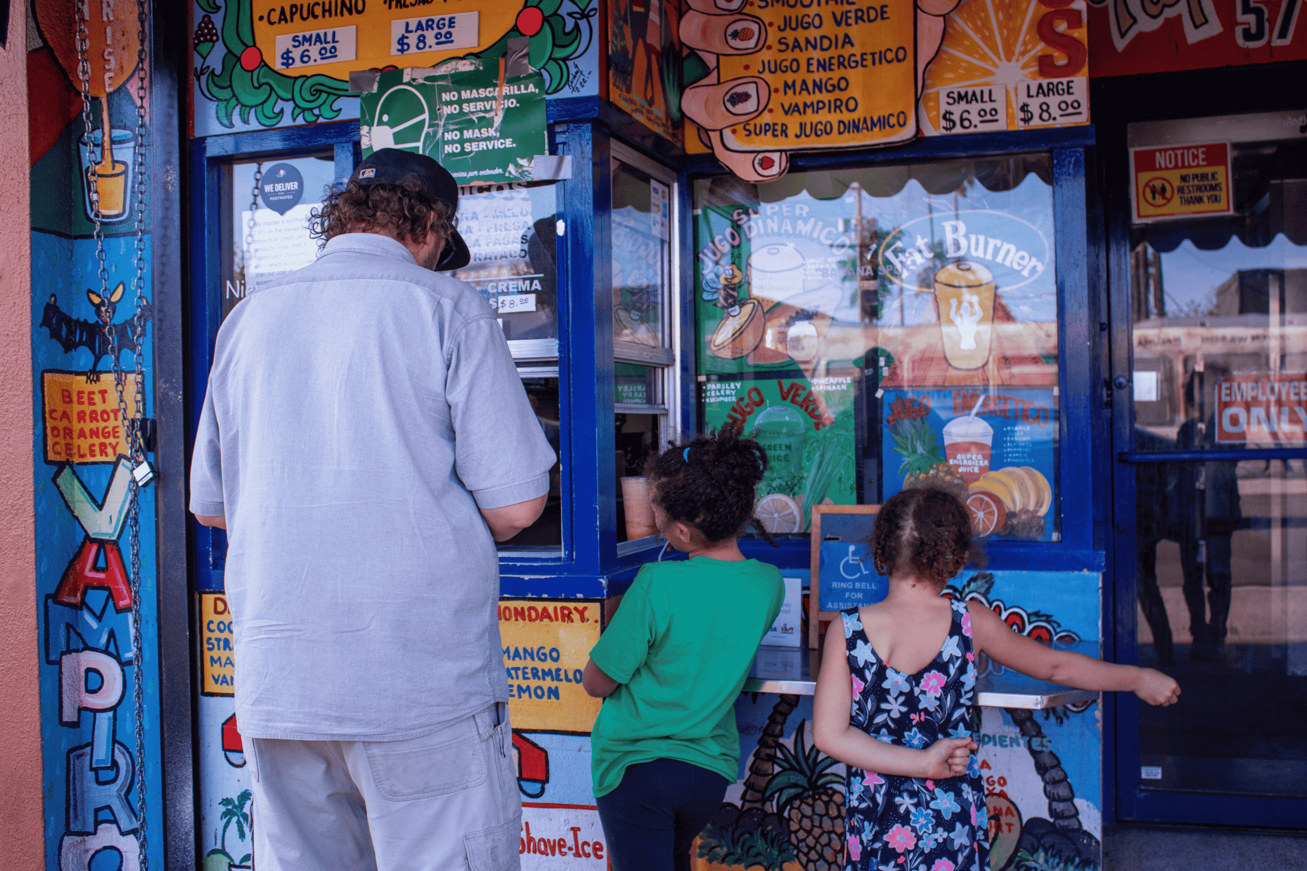 A father pays for his two young daughters, excited behind him, at a juice and smoothie stand that's bright and colorful [mostly blues and oranges].