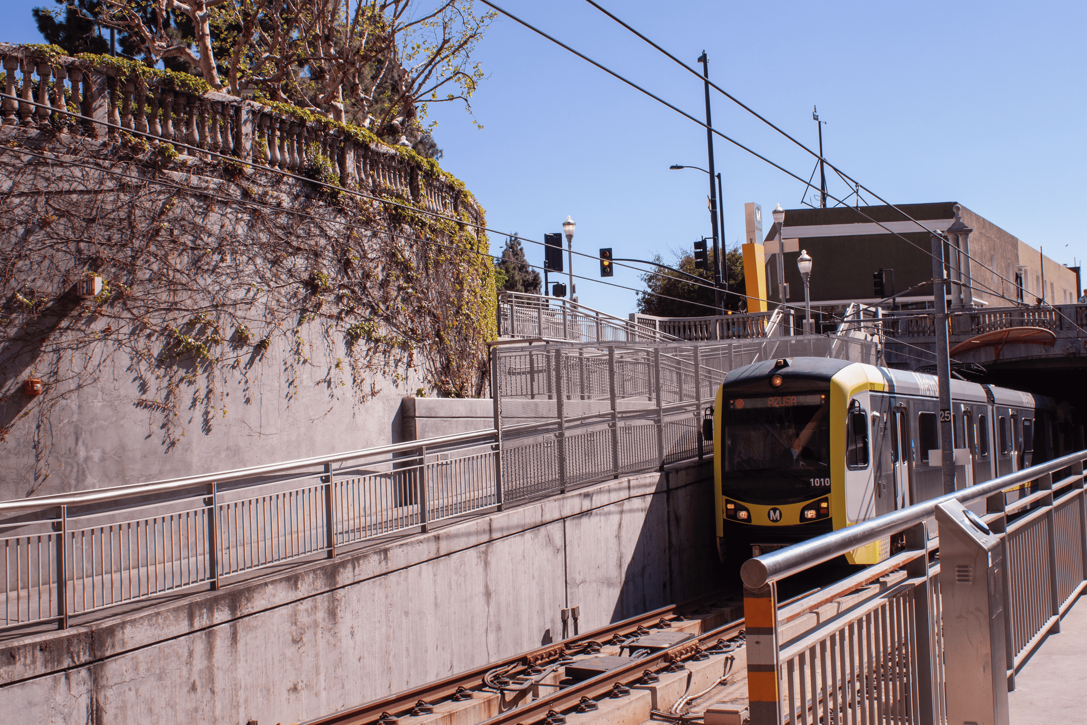 A yellow train pulls into a fairly gray and concrete station from inside a tunnel. Vines creep up one wall of the station, and a bright blue sky is visible behind it all.