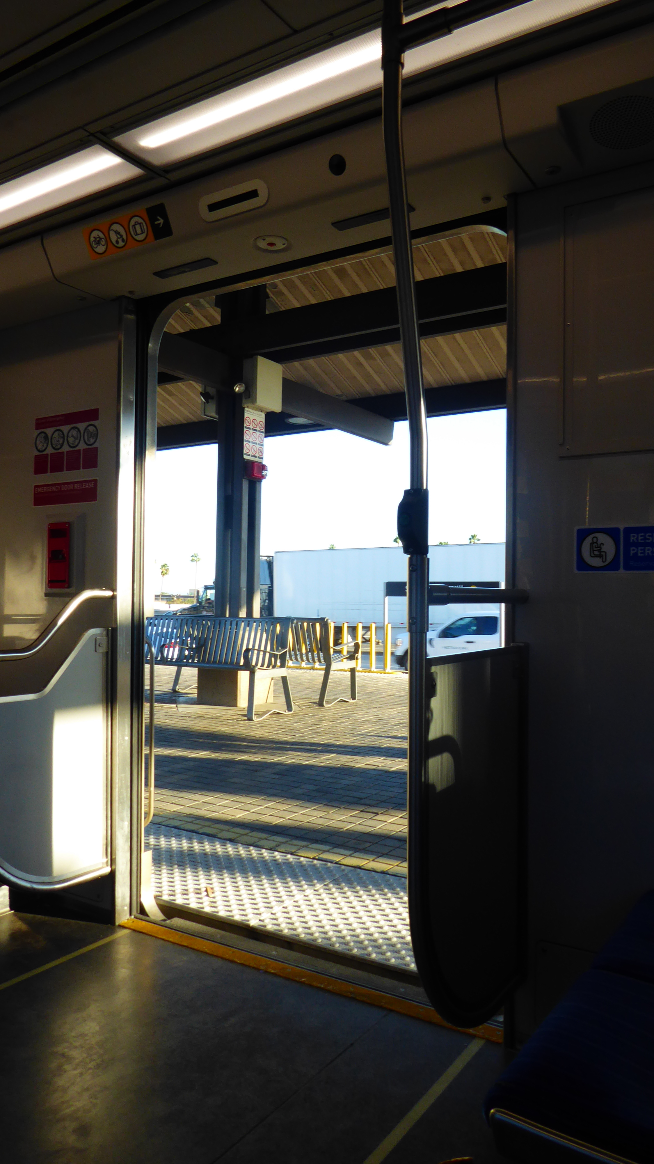 A photo of an open train door-- the inside is dark and warm colored, with a yellow light streaming in. You can see a bench and a truck passing by through the door.